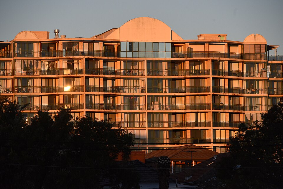 An eight-storey apartment block with rows of balconies lit orange by the setting sun, above the red tiled roofs of older houses.