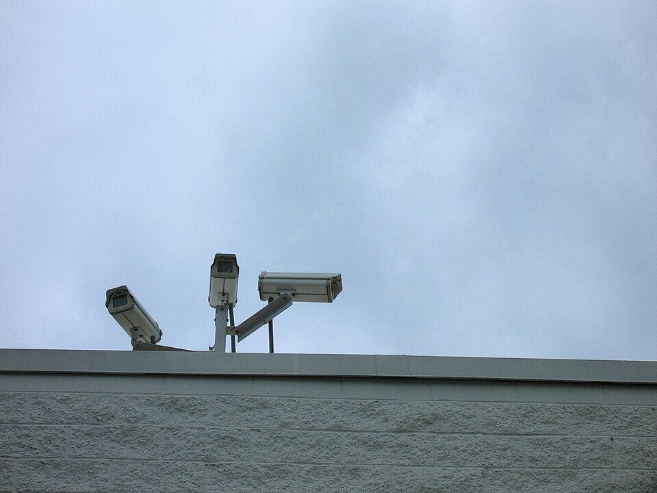 Three CCTV cameras on short poles on the parapet of a block building, pointing in different directions against a grey sky.
