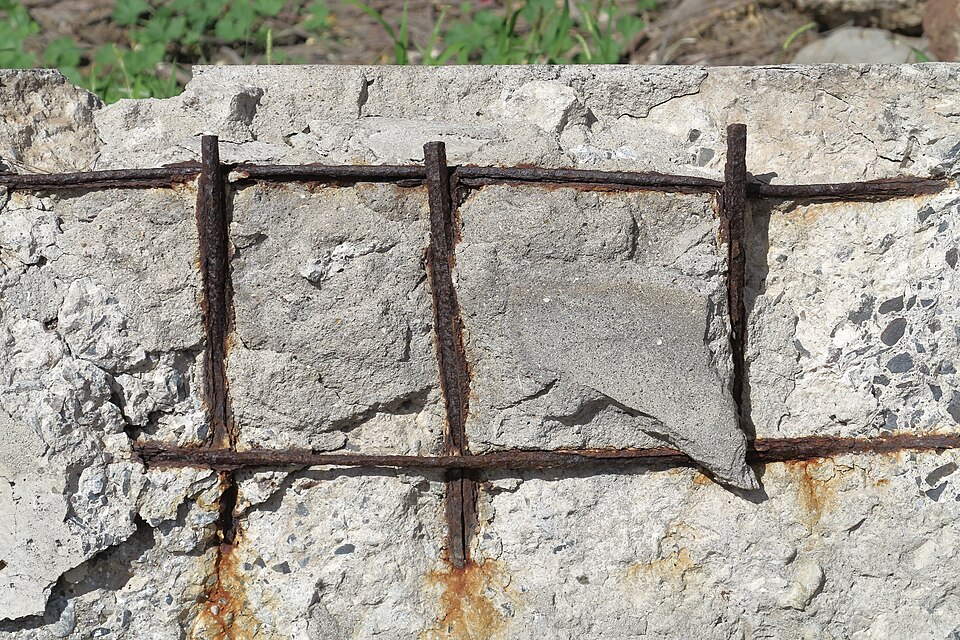 A broken concrete slab edge where the surface has fallen away, exposing a grid of rusted steel reinforcing bars with orange rust stains on the concrete below.
