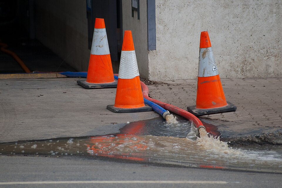 Red and blue pump hoses running out of an underground car park entrance past orange traffic cones, discharging muddy water into the gutter.