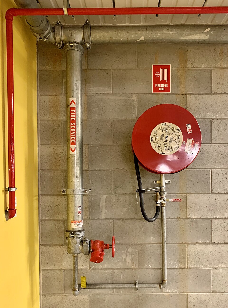 A red fire hose reel with its sign on a block wall, beside a galvanised fire service pipe with a red hydrant valve at the bottom, in a Brisbane building.