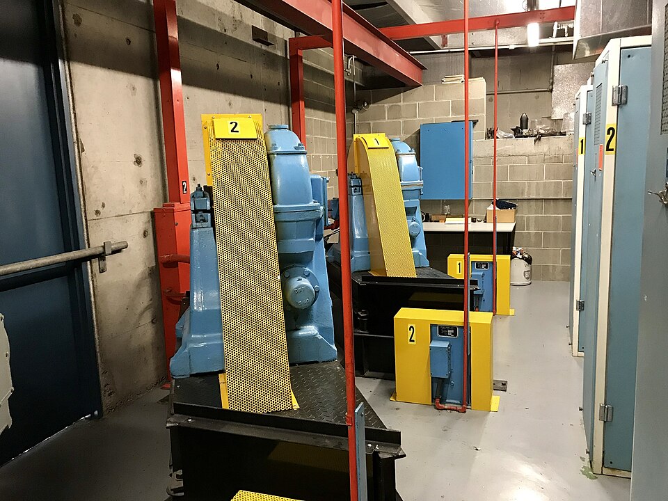 A lift motor room with two blue traction machines behind yellow mesh guards, numbered 1 and 2, with red steel framing and control cabinets along the wall.