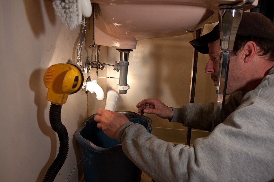 A plumber crouched under a bathroom basin, holding a section of white waste pipe over a bucket, with a work light clipped to the wall.