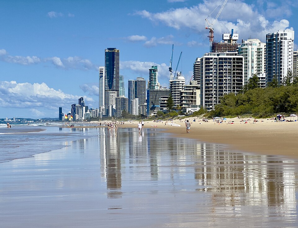 A wide beach at Surfers Paradise with apartment towers along the shore, reflected in wet sand, and cranes on new buildings behind.